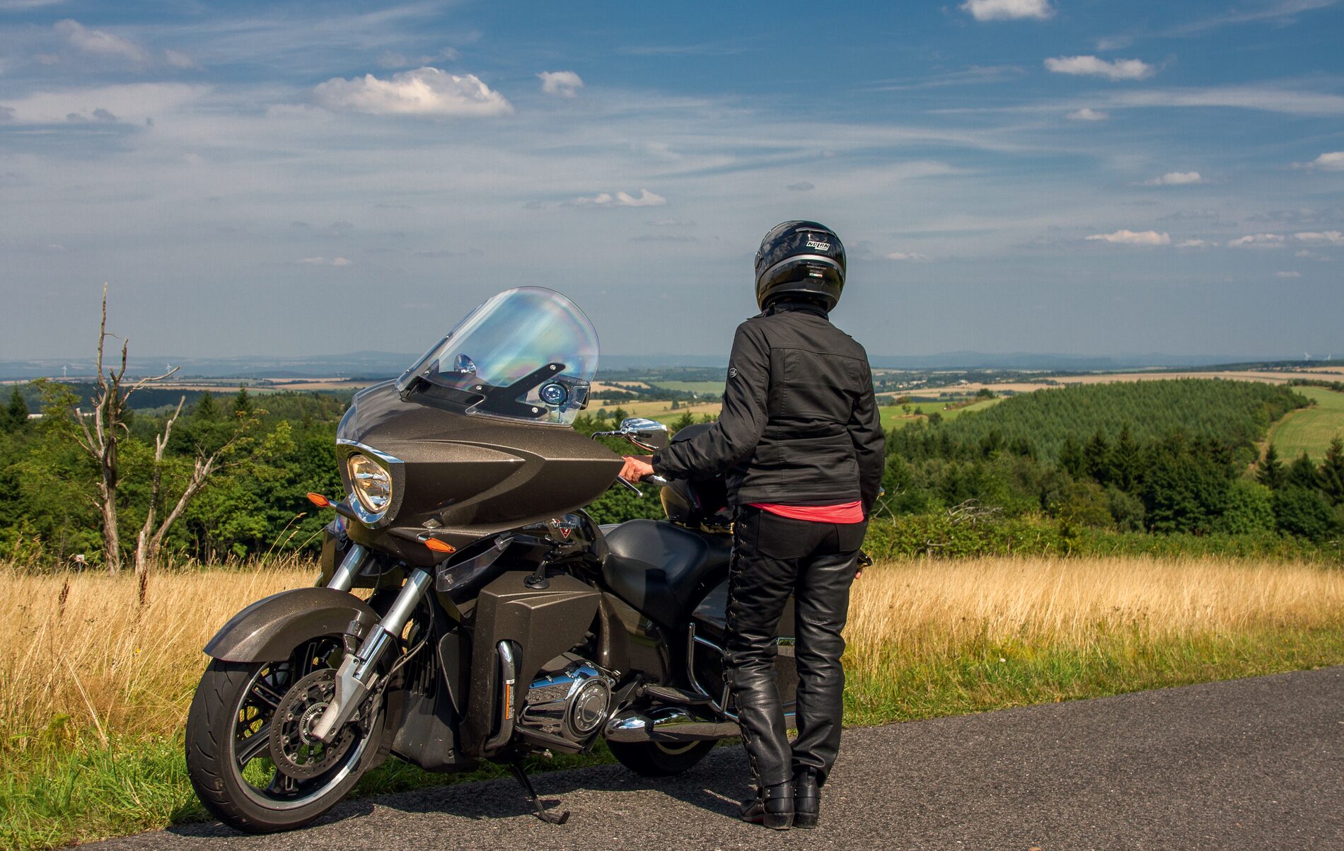 Motorradfahrerin neben ihrer Maschine an einem Straßenrand mit weitem Blick über Wiesen und Wälder Motorradfahrerin neben ihrer Maschine an einem Straßenrand mit weitem Blick über Wiesen und Wälder